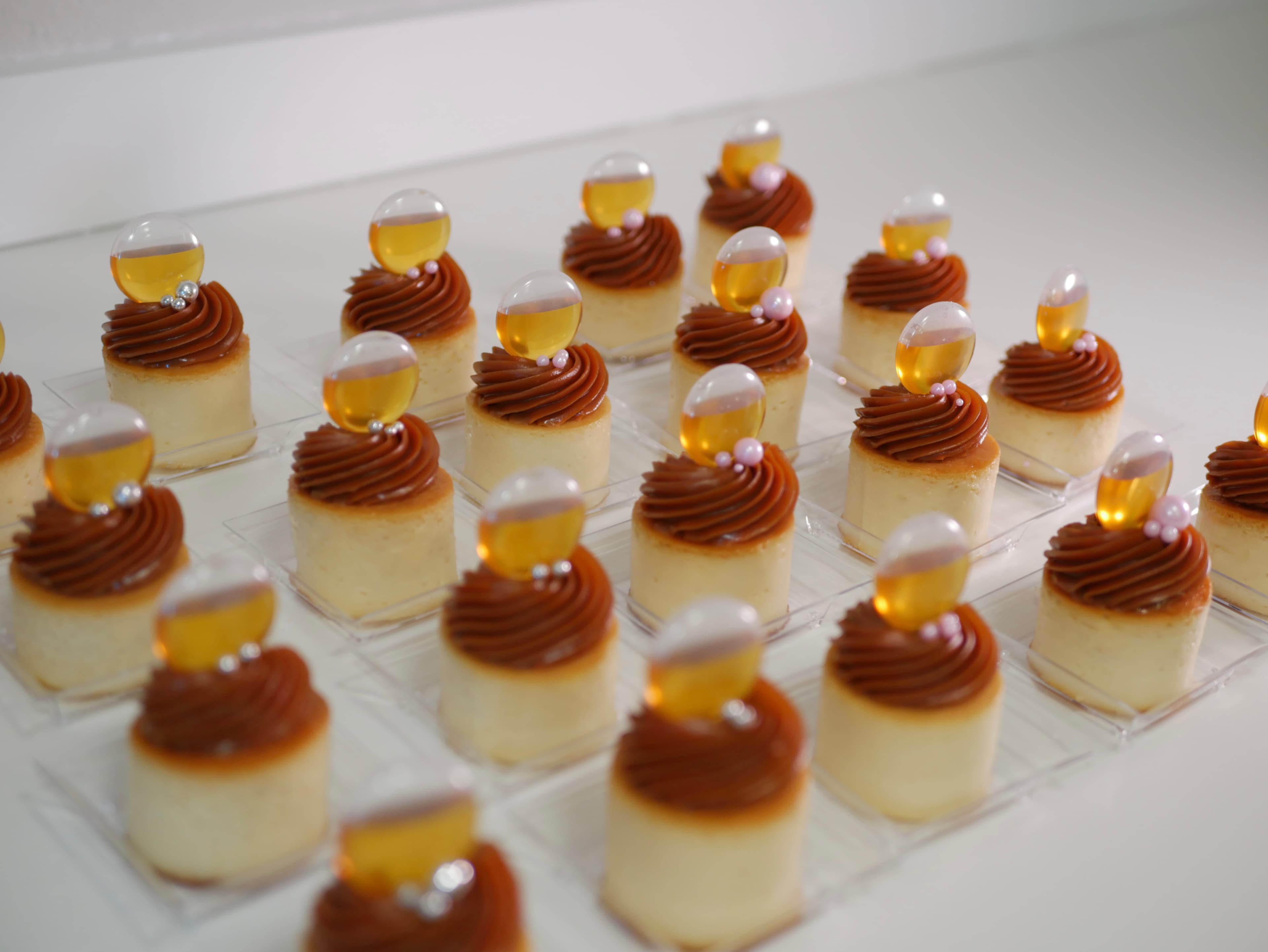Miniature cheesecakes topped with caramel swirls, silver pearls, and amber liquid-filled pipettes in rows.
